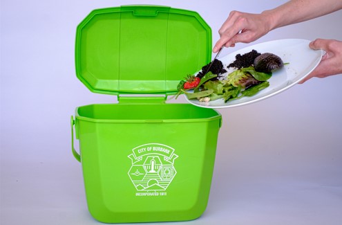 A pair of hands scraping salad food scraps off of a plate and into a green food scraps bin using a fork.