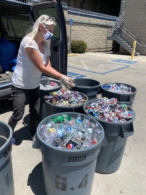 A woman standing next to several large trash bins filled with aluminum cans and plastic bottles; she is currently depositing a crushed aluminum can into a bin filled to the brim with crushed aluminum cans.